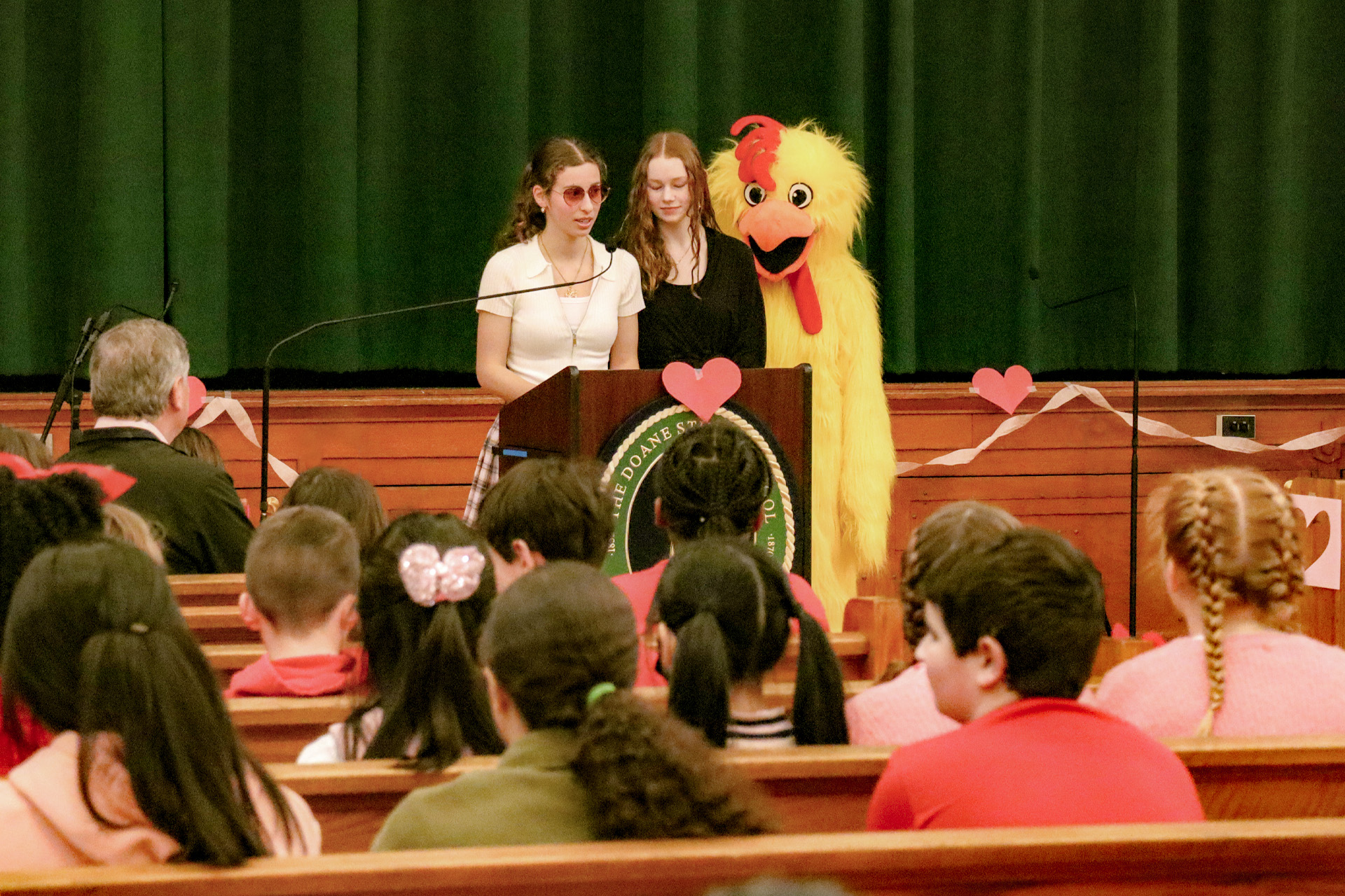 Two Doane Stuart Upper School Students address their classmates from a podium with the Thunder Chicken behind them